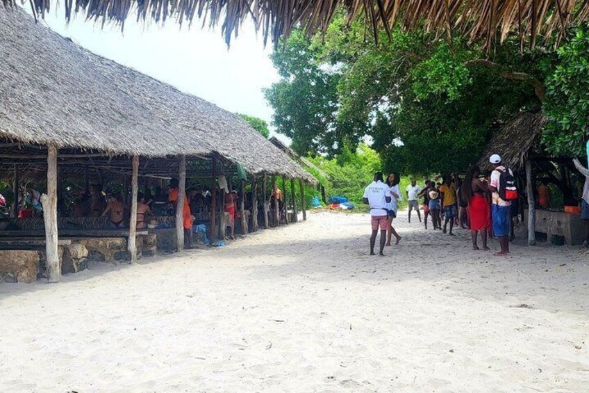 A sandy street with thatched-roof structures, people walking, and a beach in the background. 5