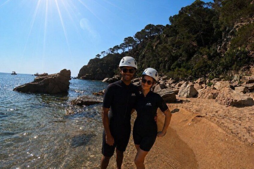 Two people standing on a rocky beach with clear water and a rocky cliff in the background. 3