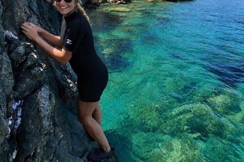 A person in a wetsuit leaning on a rocky cliff by clear blue water. 4