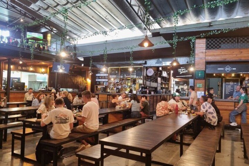 A bustling indoor dining area with wooden tables and benches, a bar in the background, and hanging greenery. 3