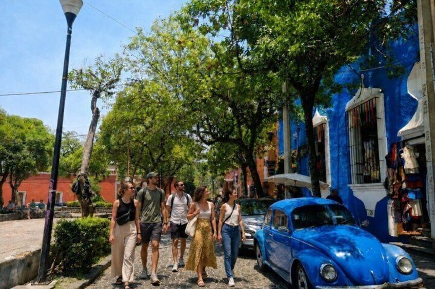 A blue car parked on a street with pedestrians and trees. 1