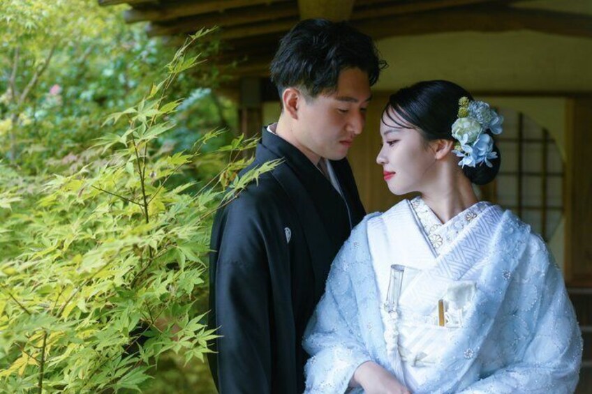 A couple in traditional Japanese attire, a man in black and a woman in white, standing in a garden with green foliage and a wooden structure in the background. 1