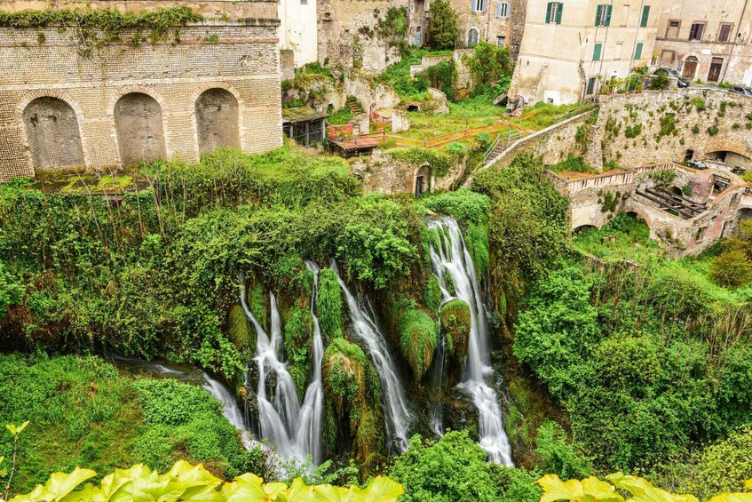 A lush waterfall cascading down a rocky cliff surrounded by dense greenery and historic buildings. 4