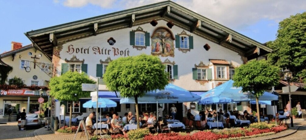 A building with 'Hotel Alte Post' signage, outdoor seating with umbrellas, and a well-maintained flower bed. 5