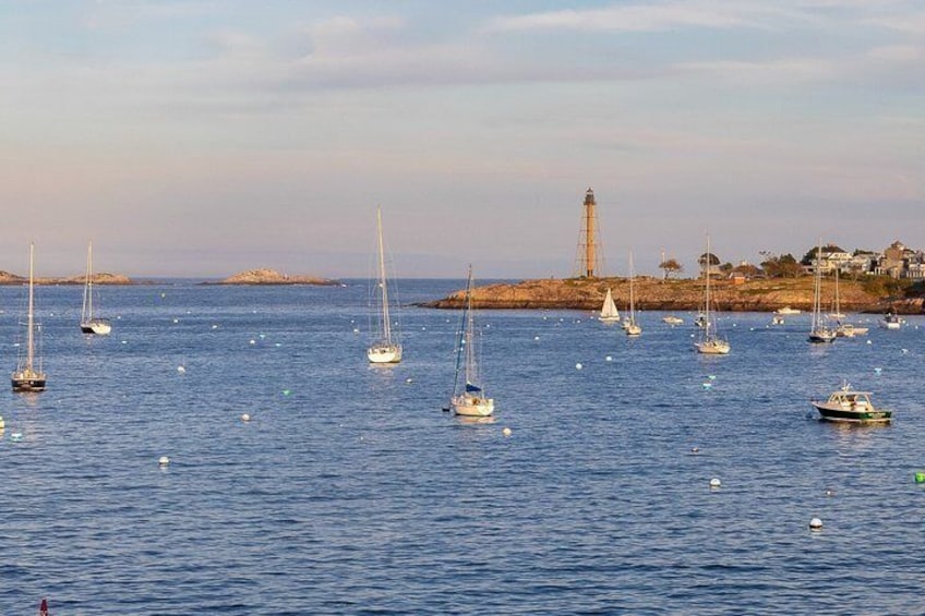 A coastal scene with multiple sailboats anchored in calm waters, a lighthouse, and rocky outcrops in the background. 4