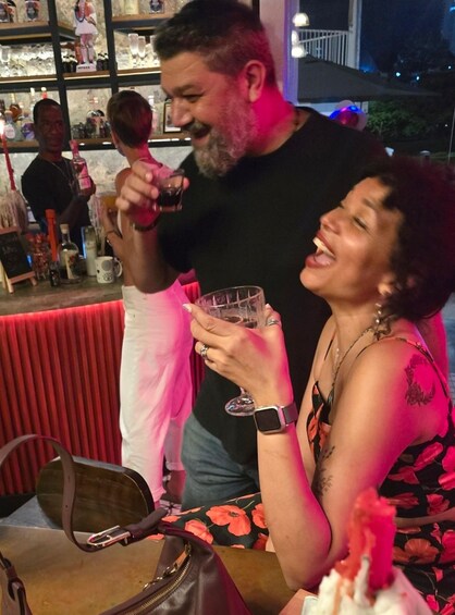 A person holding a glass, another person sitting at a table with a handbag, and a bar in the background. 4