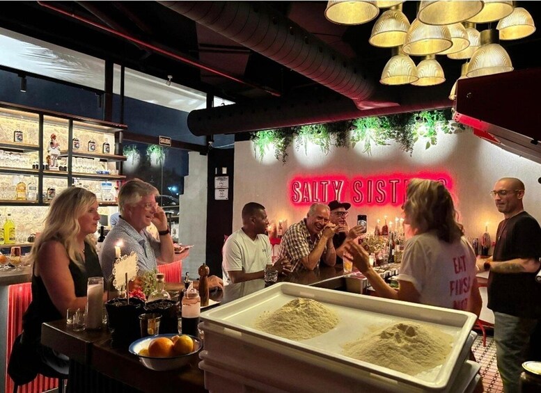 A bar with a neon sign reading 'SALTY SIST' and people socializing around tables with drinks and snacks. 2