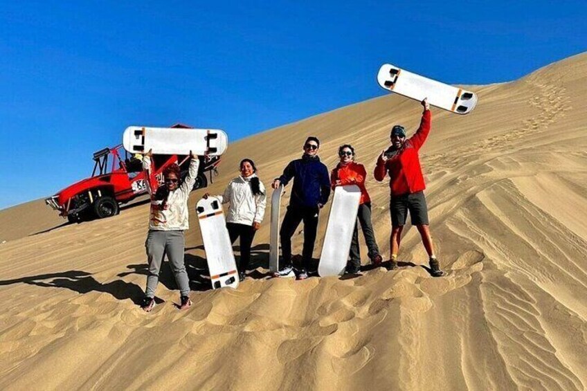 A group of people holding snowboards on a sandy dune with a red vehicle in the background. 2