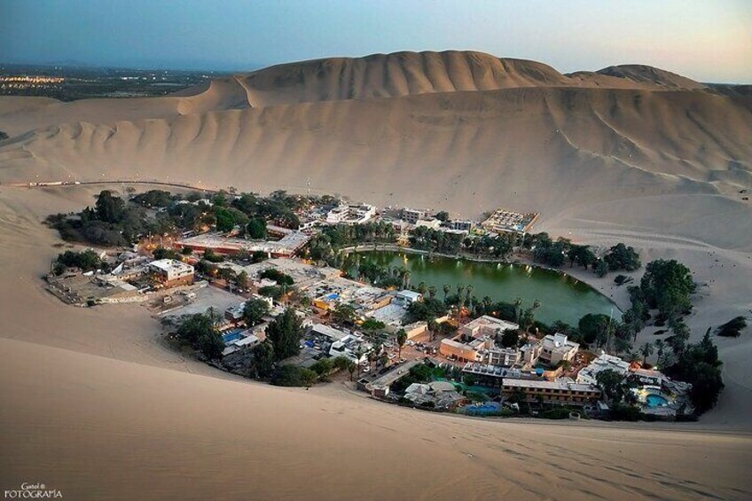 A desert landscape with sand dunes and a small settlement with buildings and a body of water. 4
