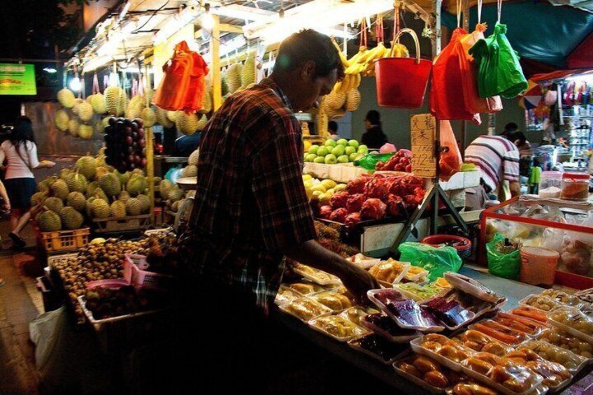 A market stall with durians and other fruits, a man at the counter, and hanging baskets of bananas. 3