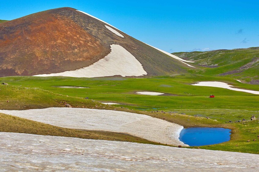 A mountain with snow patches, a blue pond, and a red vehicle in a green valley. 5