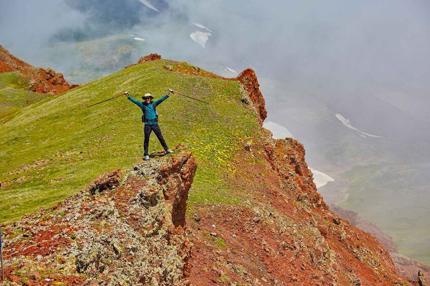 A person standing on a mountain peak with arms raised, surrounded by rocky terrain and patches of snow. 2