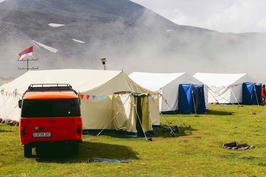 A red van parked in front of tents with a flag on a mountain. 4