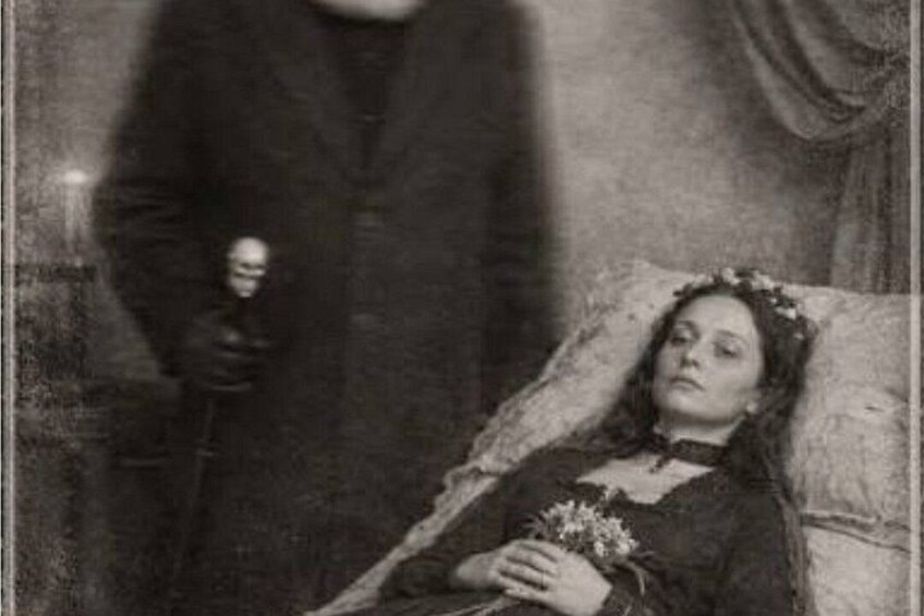 A person in a dark coat holding a bottle, standing next to a reclining individual holding a bouquet. 2