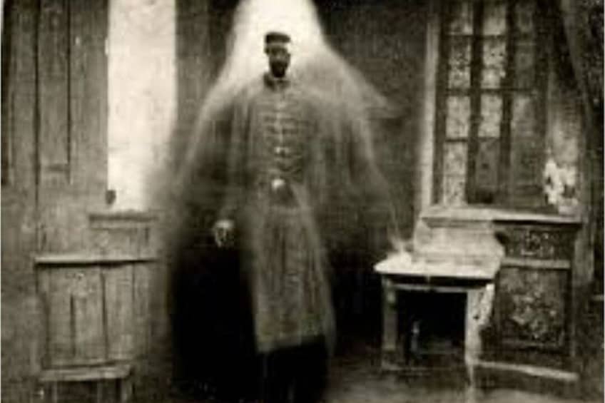 A person in a long coat standing in a room with wooden furniture and a window. 4