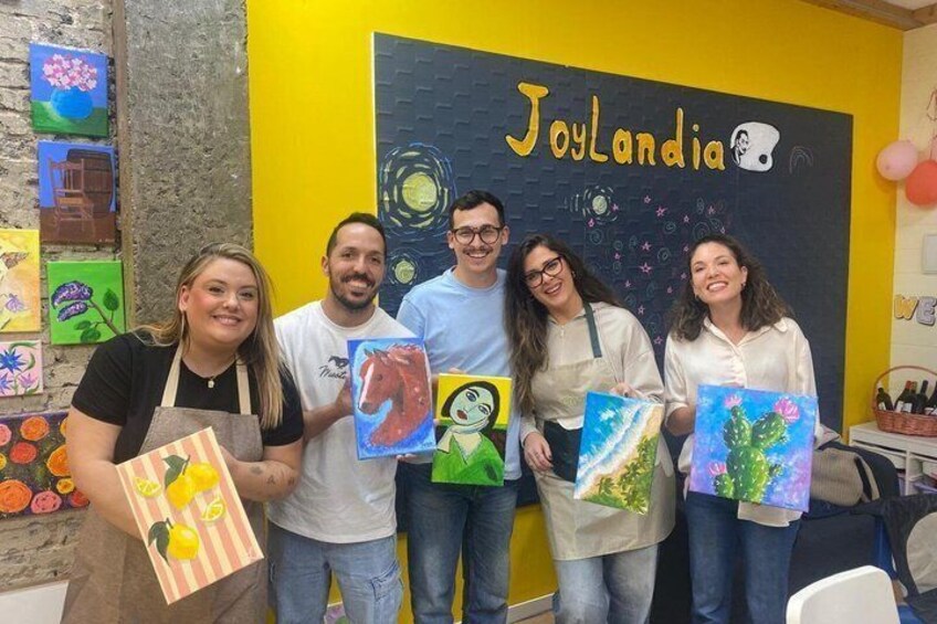 A group of people holding painted canvases in front of a wall with the text 'Joylandia' and a chalkboard with drawings. 5