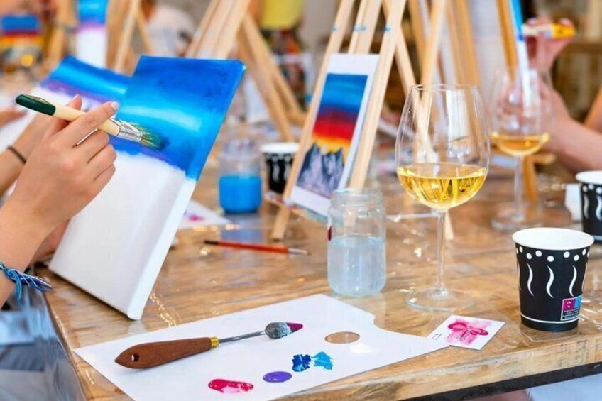 An artist painting a landscape on canvas, with paint jars, brushes, and a wine glass on a table. 4