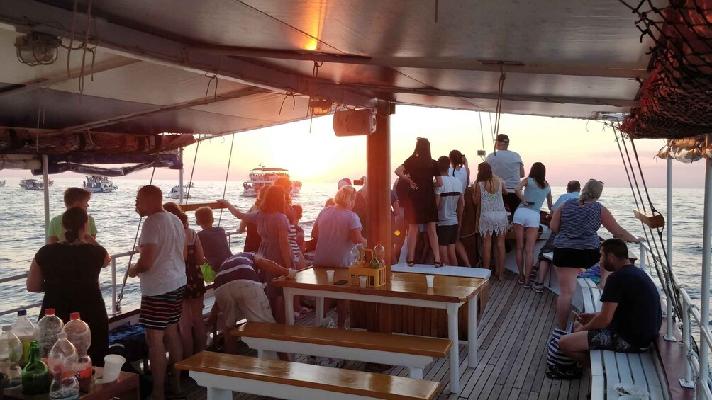 A group of people on a boat deck during sunset, with the ocean and other boats in the background. 3