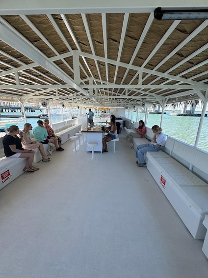 A covered boat deck with people seated and standing, a central table, and a view of the water. 2