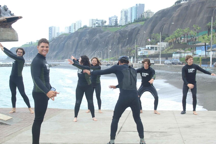 A group of people in wetsuits performing a synchronized dance on a concrete stage by the beach with a rocky cliff and buildings in the background. 4
