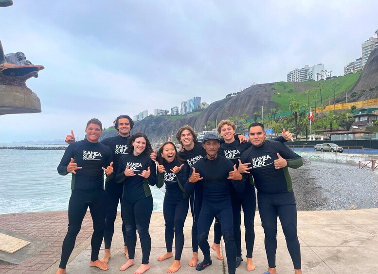 A group of people in wetsuits by a rocky coastline with buildings in the background. 1