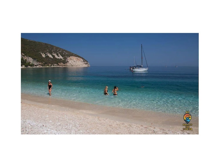 A beach with clear turquoise water, a sailboat, and rocky hills in the background. 4
