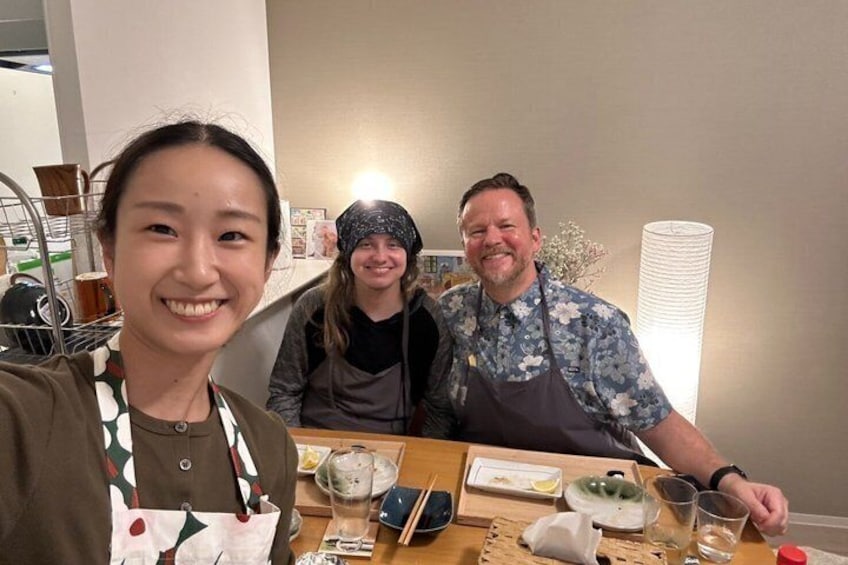 Three people seated at a table with food and drinks, in a room with a lamp and a shelf in the background. 5