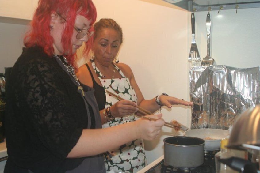 Two people cooking in a kitchen with a pot on the stove and a frying pan on the counter. 2