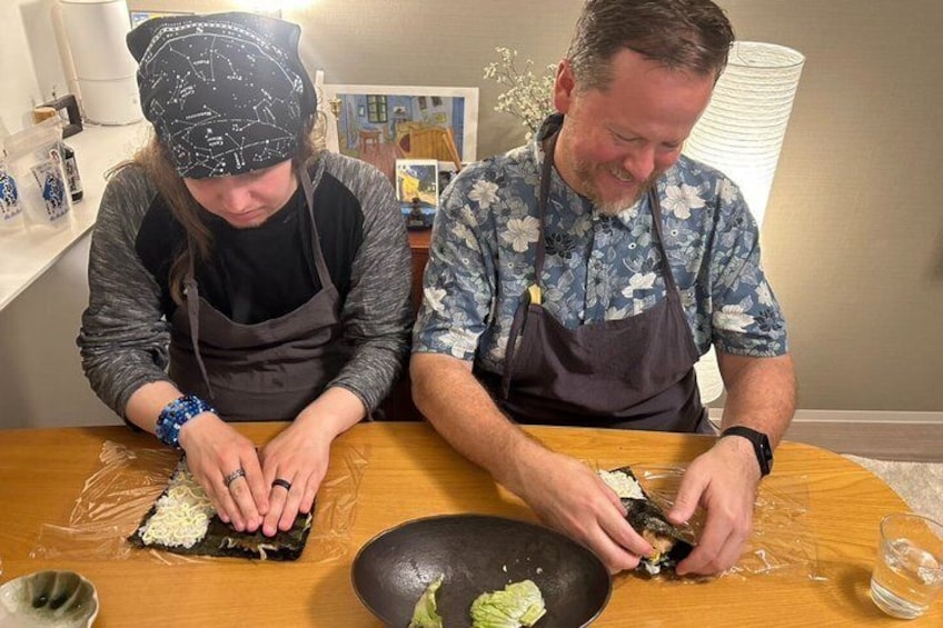 Two people preparing food, one wearing a bandana and apron, and the other in a floral shirt and apron, with a bowl and plastic wrap on the table. 4
