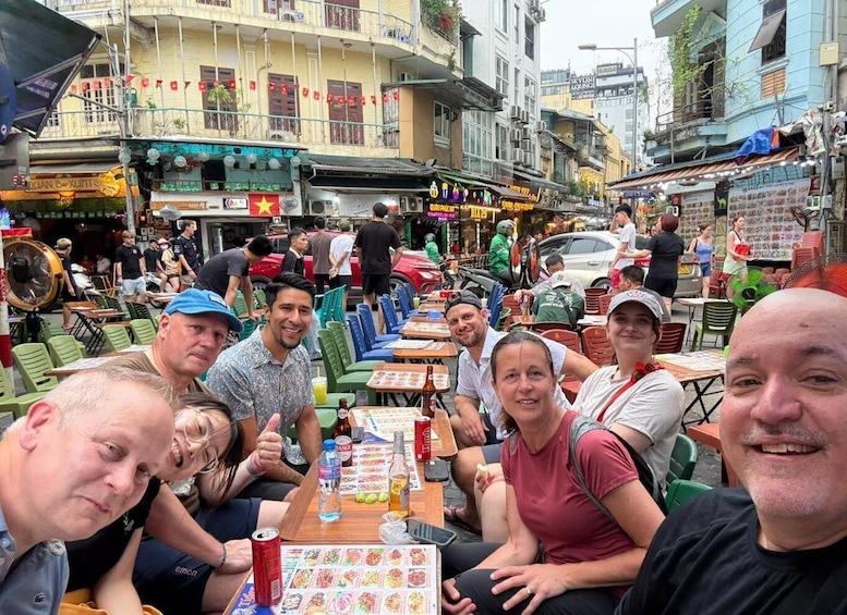 A group of people seated at an outdoor table with a variety of food items and drinks, surrounded by urban buildings and a busy street scene. 1