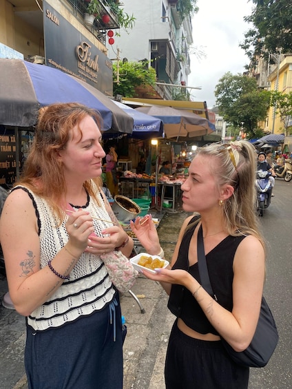 Two women eating street food in an urban setting with market stalls and a salon in the background. 5