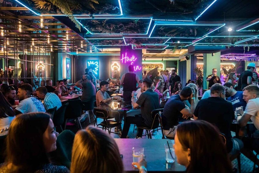 A crowded indoor bar with neon signage and people socializing. 1