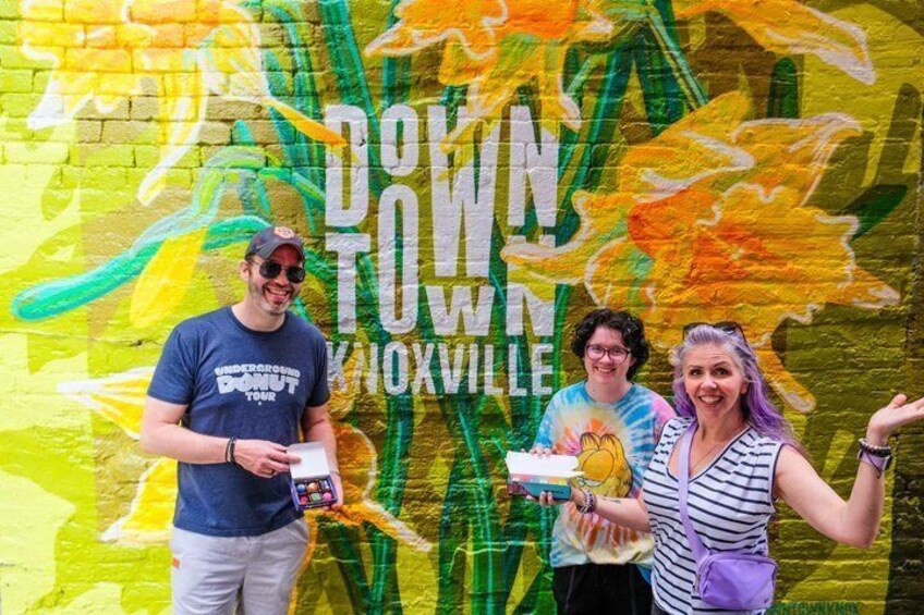 Three people standing in front of a colorful mural with 'DOWNTOWN KNOXVILLE' text. 1