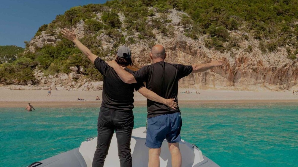 Two people on a boat facing a rocky coastline with a beach in the background. 3