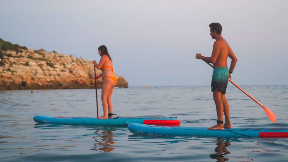 Two people paddleboarding on calm water with a rocky coastline in the background. 1