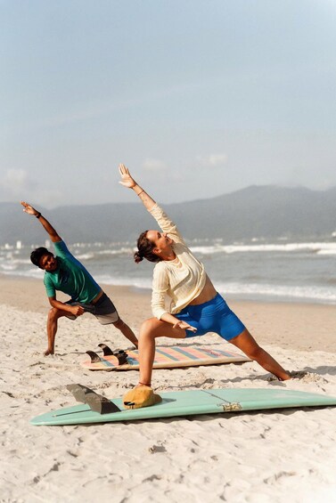 Two people practicing yoga on a beach with surfboards and ocean in the background. 4
