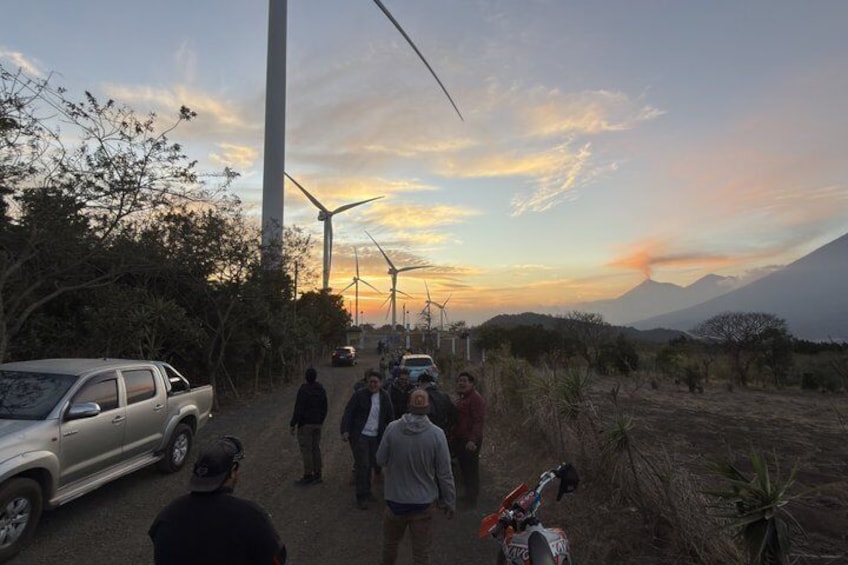 A group of people standing near wind turbines, a parked car, and a motorcycle in a dry landscape with a mountain in the background. 3
