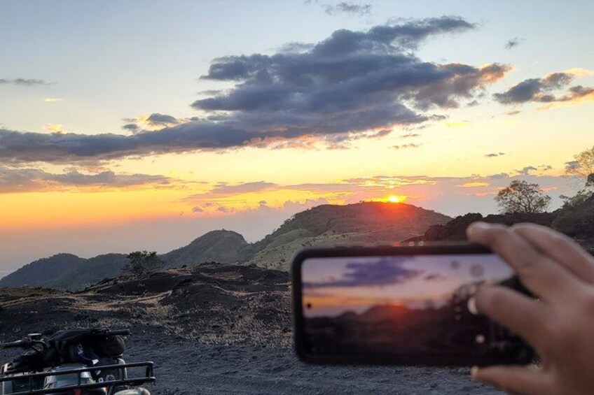 Una persona fotografiando un atardecer con un teléfono inteligente, un vehículo y un paisaje montañoso. 5