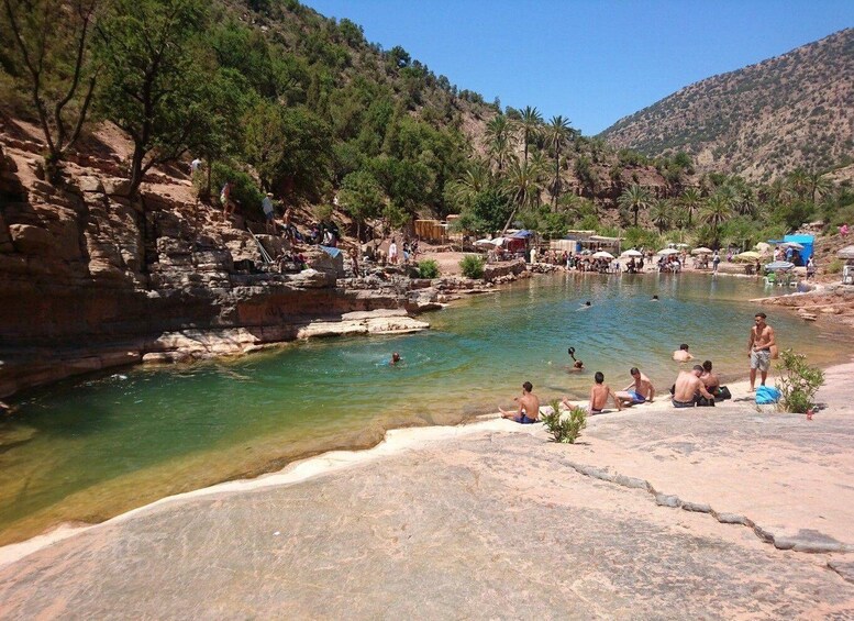 A natural pool surrounded by rocky terrain and sparse vegetation, with people swimming and relaxing on the shore. 3
