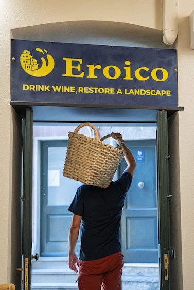 A person carrying a woven basket over their head, approaching a door with a sign that reads 'Eroico - Drink Wine, Restore a Landscape.' 3