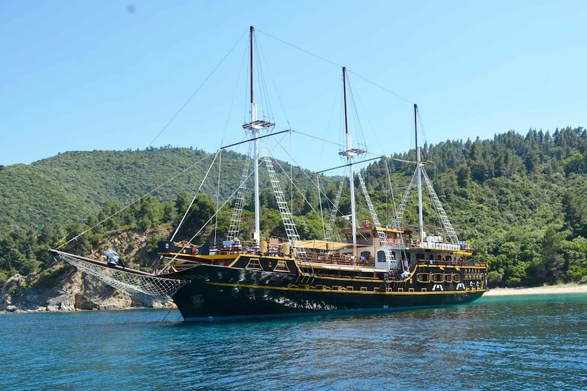A large sailing ship with multiple masts and a deck full of people, anchored near a forested coastline. 1