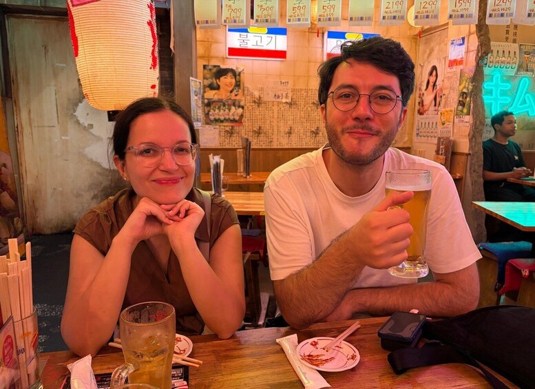 Two people sitting at a table in a restaurant with various items on the table, including a glass of beer and chopsticks. 3
