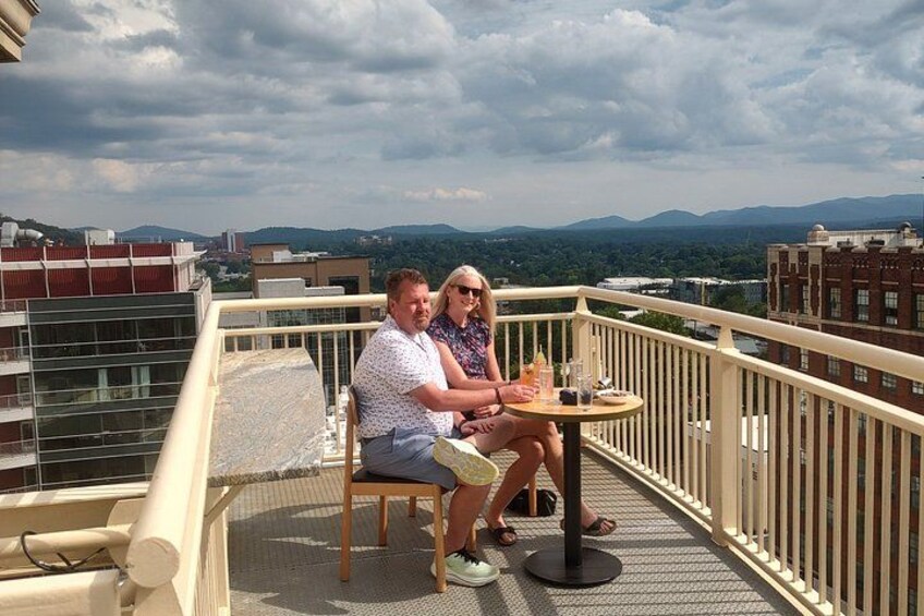 Una coppia seduta su un balcone con un tavolo, con vista su un paesaggio urbano che include edifici e montagne all'orizzonte. 3