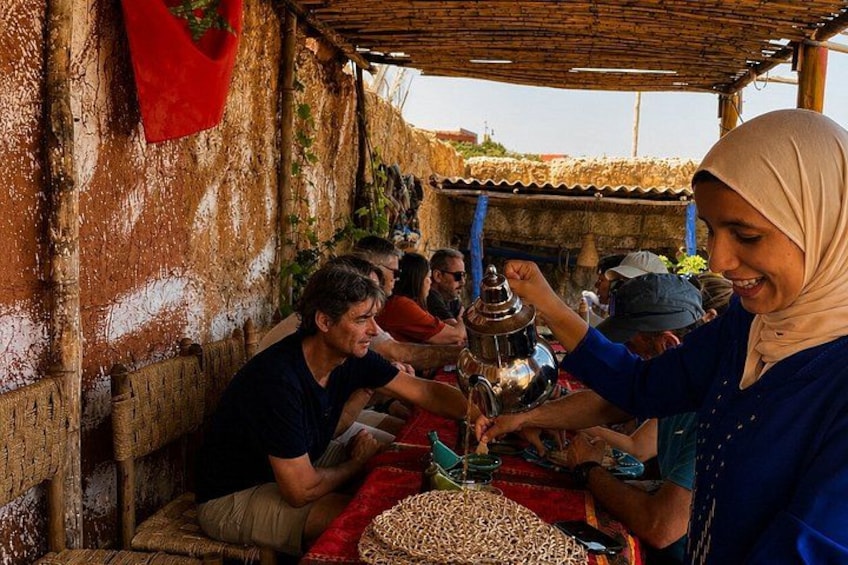 A group of people engaged in a traditional activity, possibly a cultural or religious ceremony, in a rustic outdoor setting with a thatched roof and woven furniture. 3