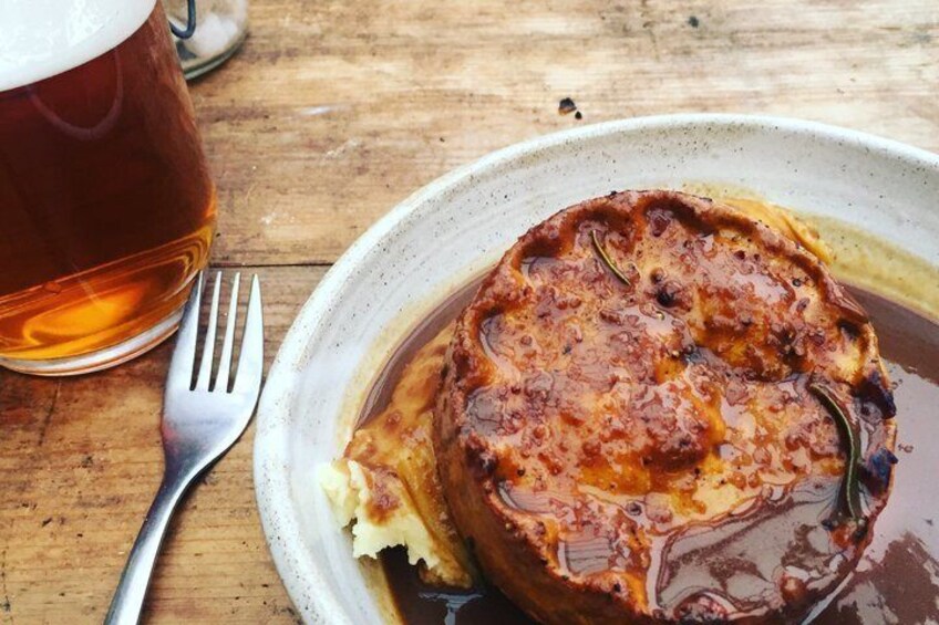 A plate of food with a liquid gravy, a fork, and a glass of beer on a wooden table. 5