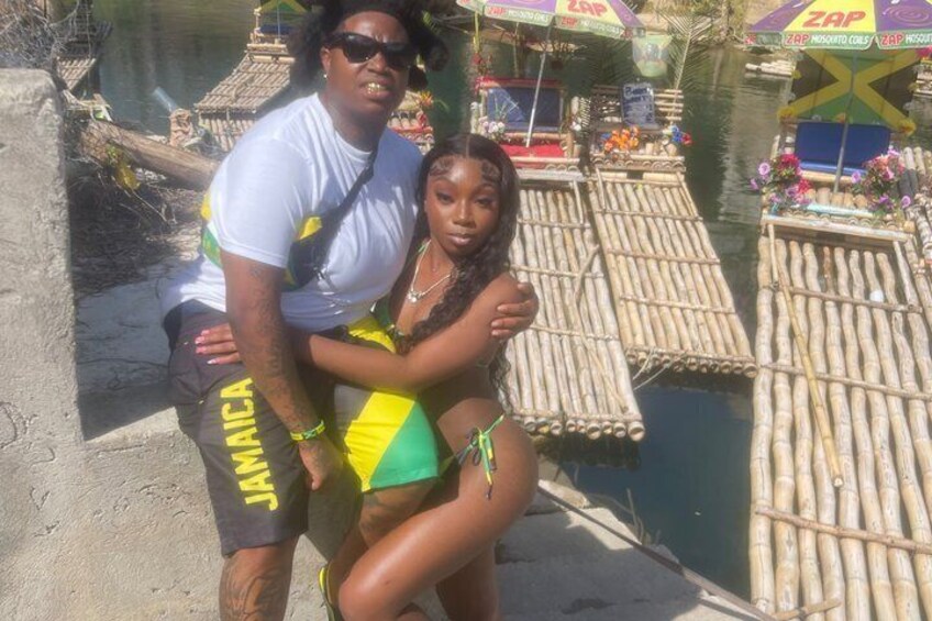 A person in a white shirt and black shorts with 'JAMAICA' on the side, standing next to a person in a green and yellow swimsuit. In the background, there are bamboo rafts with 'ZAP' branding and a flag with a green and yellow design. 3