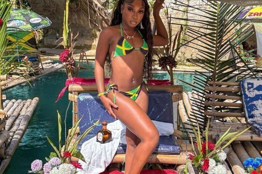 A person in a green and yellow bikini sitting on a lounge chair by a pool with tropical plants and a drink. 2