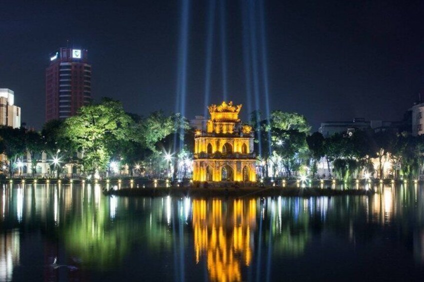 A well-lit historic building with a reflection in the water, surrounded by trees and modern buildings. 1