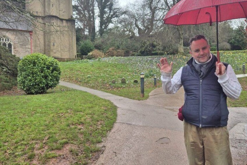 A person holding a red umbrella, standing on a paved path with a stone building and a green bush in the background. 1