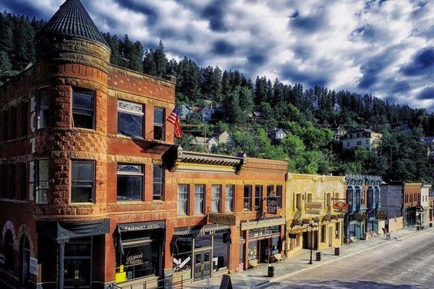 A historic street with brick buildings, a prominent corner building with a tower, and a forested hill in the background. 4